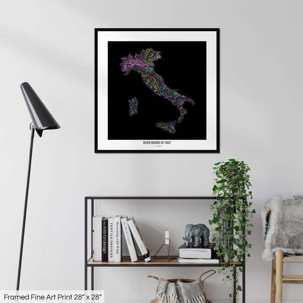 A colourful Grasshopper Geography map on a white wall, home decor in a living room. The map is in a black frame with a thick white mat. The title of the map is printed on the mat. The map hangs above a black shelf with plants and books on. A black lamp stands in the corner.