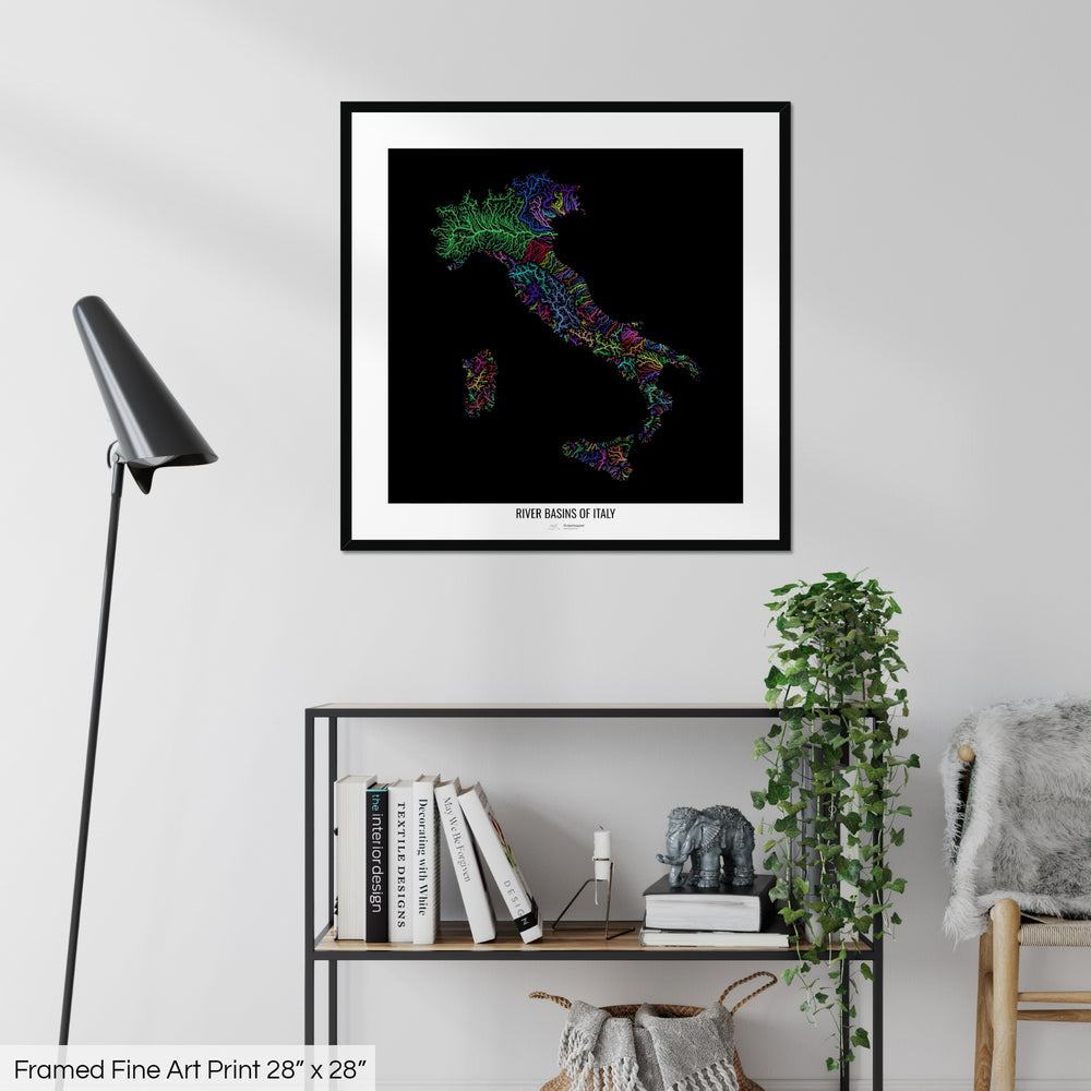 A colourful Grasshopper Geography map on a white wall, home decor in a living room. The map is in a black frame with a thick white mat. The title of the map is printed on the mat. The map hangs above a black shelf with plants and books on. A black lamp stands in the corner.