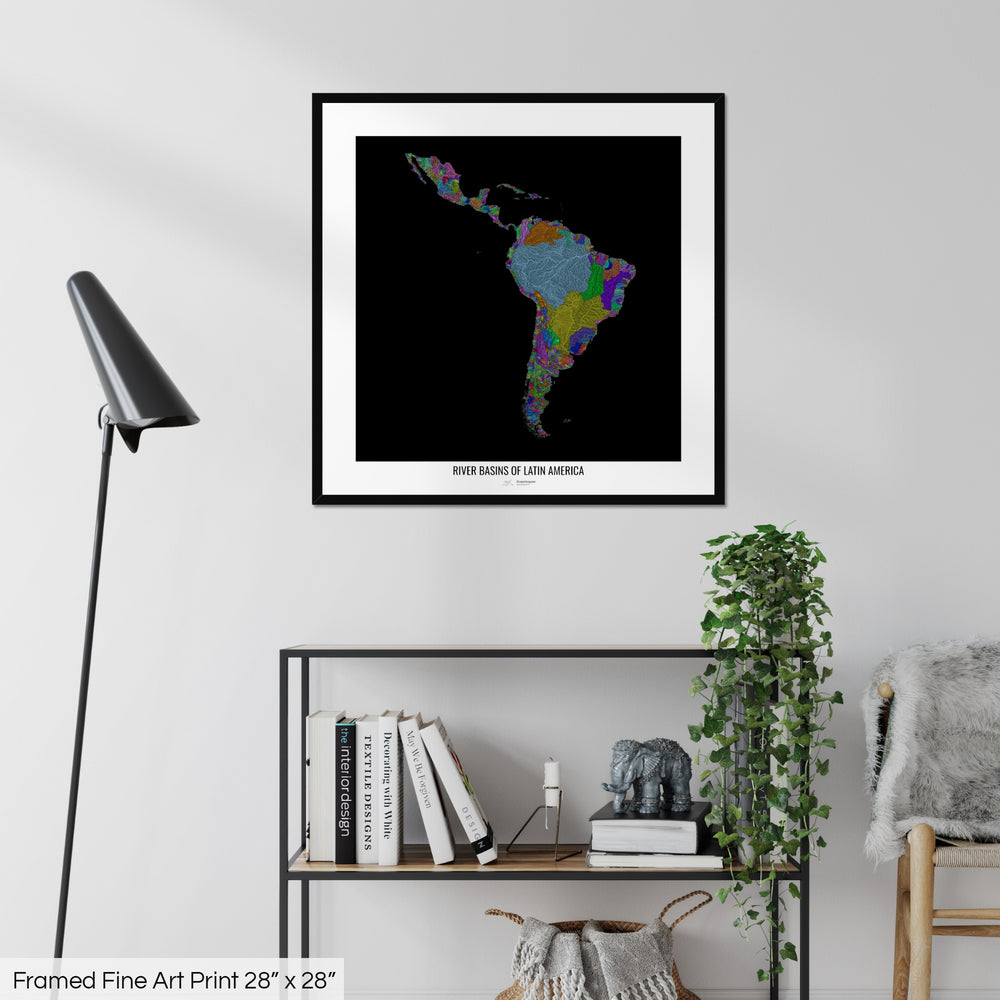 A colourful Grasshopper Geography map on a white wall, home decor in a living room. The map is in a black frame with a thick white mat. The title of the map is printed on the mat. The map hangs above a black shelf with plants and books on. A black lamp stands in the corner.