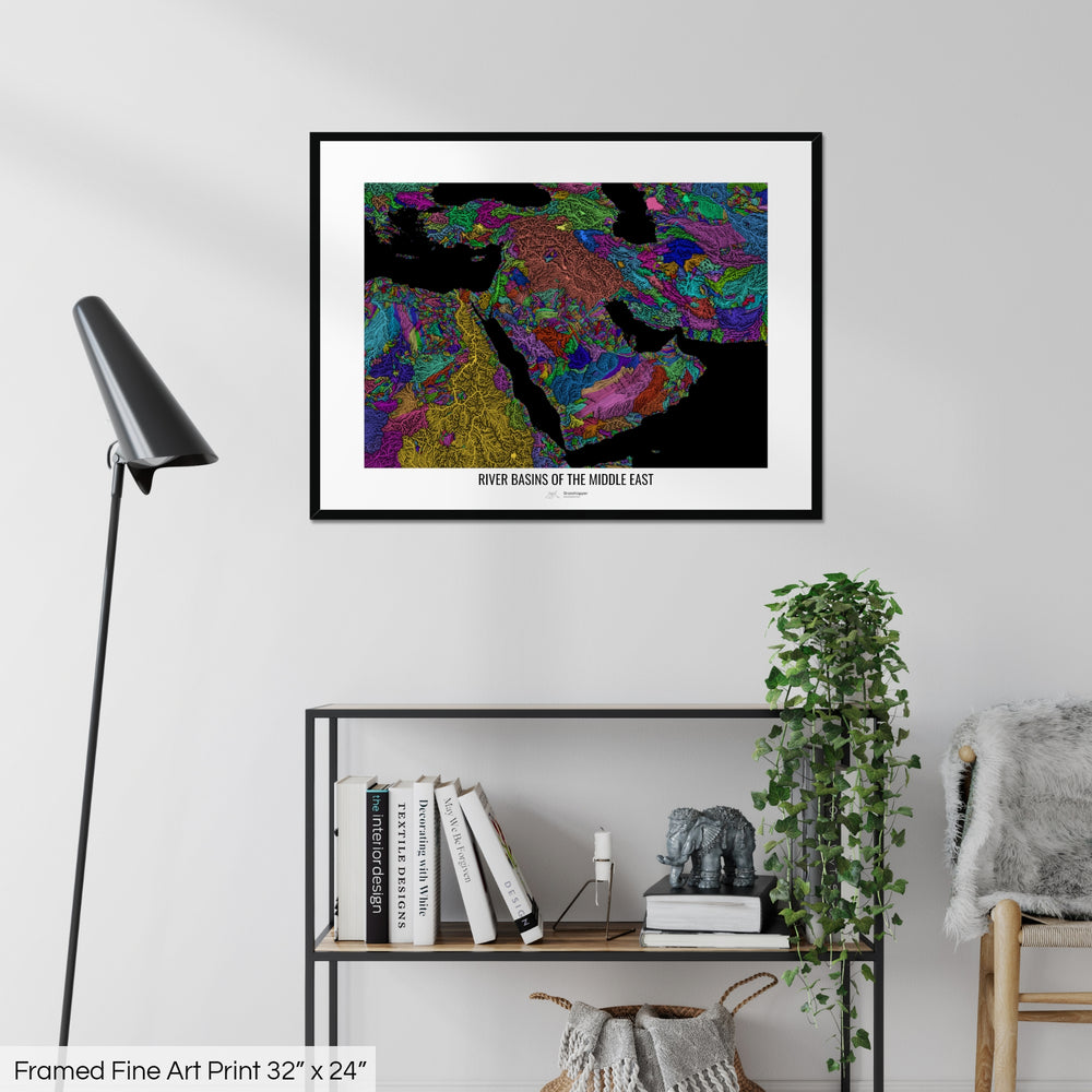 A colourful Grasshopper Geography map on a white wall, home decor in a living room. The map is in a black frame with a thick white mat. The title of the map is printed on the mat. The map hangs above a black shelf with plants and books on. A black lamp stands in the corner.