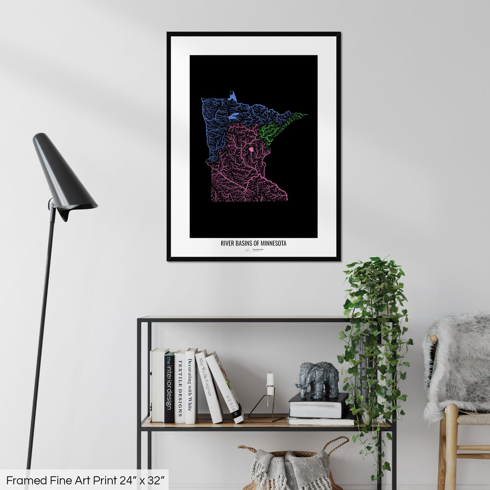 A colourful Grasshopper Geography map on a white wall, home decor in a living room. The map is in a black frame with a thick white mat. The title of the map is printed on the mat. The map hangs above a black shelf with plants and books on. A black lamp stands in the corner.