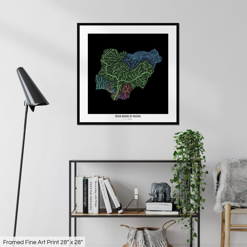 A colourful Grasshopper Geography map on a white wall, home decor in a living room. The map is in a black frame with a thick white mat. The title of the map is printed on the mat. The map hangs above a black shelf with plants and books on. A black lamp stands in the corner.
