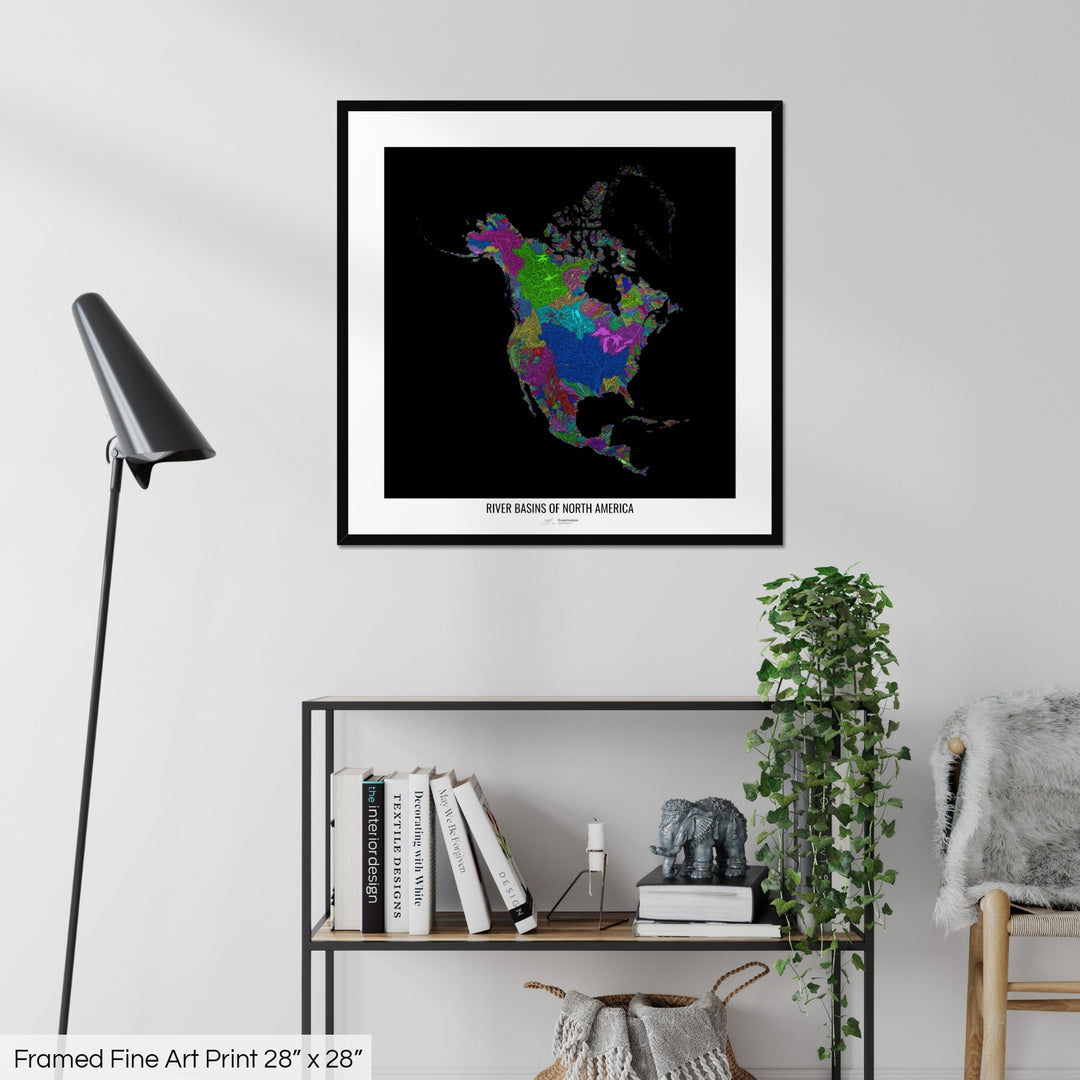 A colourful Grasshopper Geography map on a white wall, home decor in a living room. The map is in a black frame with a thick white mat. The title of the map is printed on the mat. The map hangs above a black shelf with plants and books on. A black lamp stands in the corner.