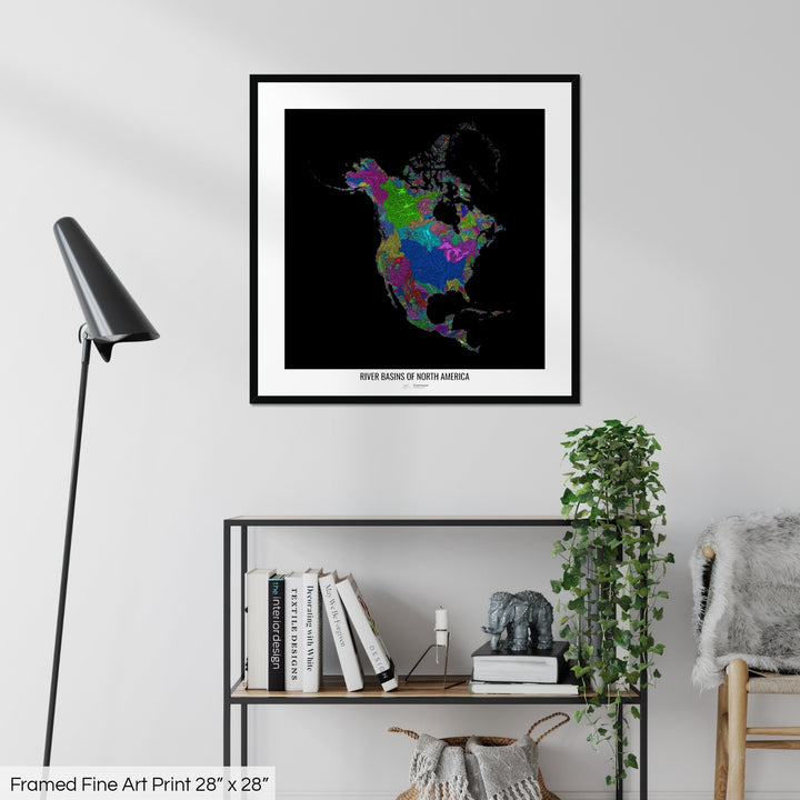 A colourful Grasshopper Geography map on a white wall, home decor in a living room. The map is in a black frame with a thick white mat. The title of the map is printed on the mat. The map hangs above a black shelf with plants and books on. A black lamp stands in the corner.