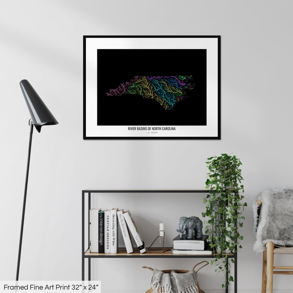 A colourful Grasshopper Geography map on a white wall, home decor in a living room. The map is in a black frame with a thick white mat. The title of the map is printed on the mat. The map hangs above a black shelf with plants and books on. A black lamp stands in the corner.