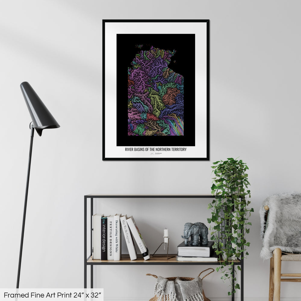 A colourful Grasshopper Geography map on a white wall, home decor in a living room. The map is in a black frame with a thick white mat. The title of the map is printed on the mat. The map hangs above a black shelf with plants and books on. A black lamp stands in the corner.