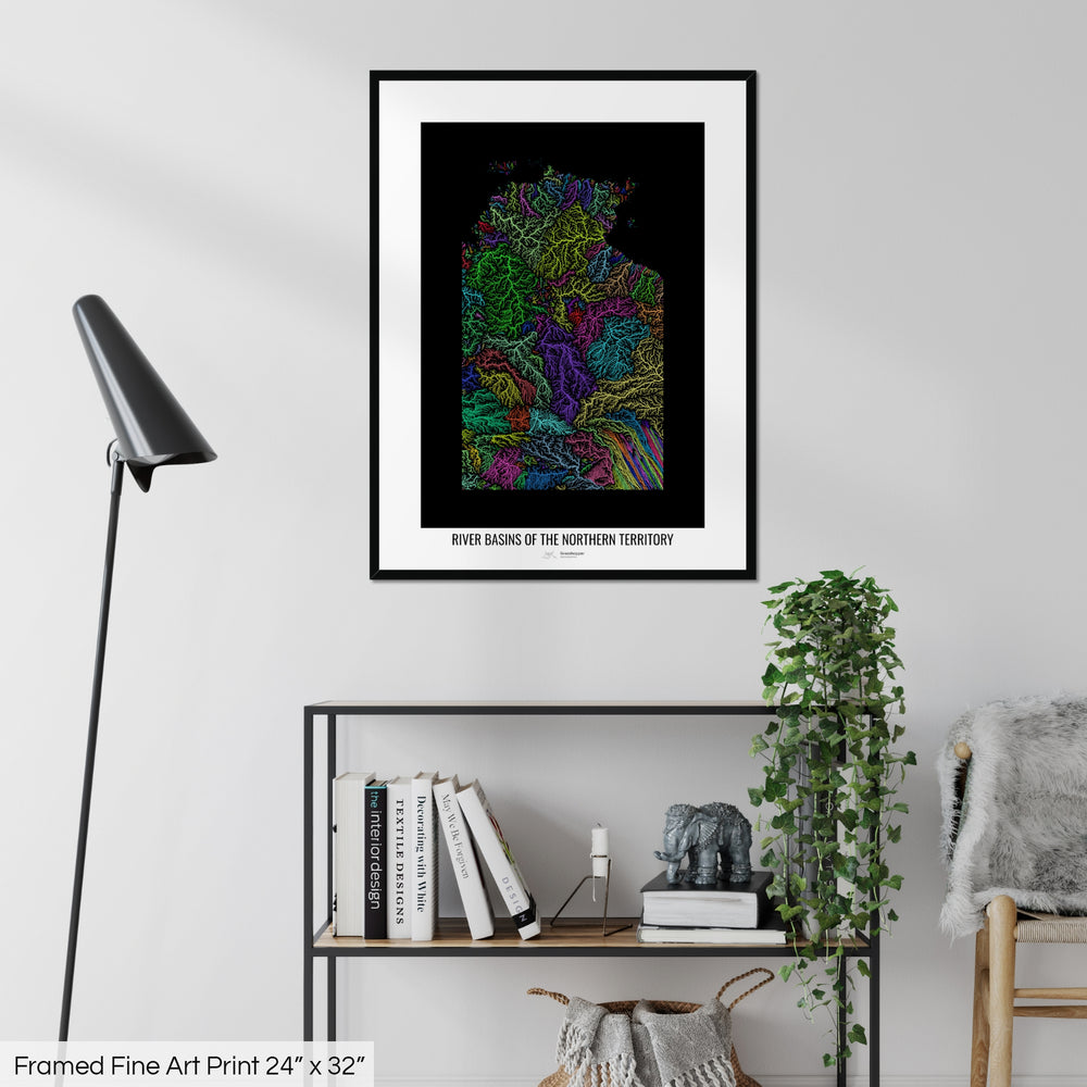 A colourful Grasshopper Geography map on a white wall, home decor in a living room. The map is in a black frame with a thick white mat. The title of the map is printed on the mat. The map hangs above a black shelf with plants and books on. A black lamp stands in the corner.