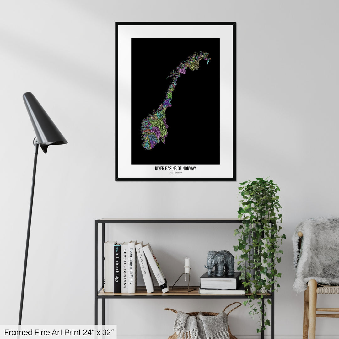 A colourful Grasshopper Geography map on a white wall, home decor in a living room. The map is in a black frame with a thick white mat. The title of the map is printed on the mat. The map hangs above a black shelf with plants and books on. A black lamp stands in the corner.