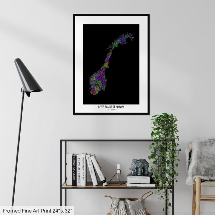 A colourful Grasshopper Geography map on a white wall, home decor in a living room. The map is in a black frame with a thick white mat. The title of the map is printed on the mat. The map hangs above a black shelf with plants and books on. A black lamp stands in the corner.