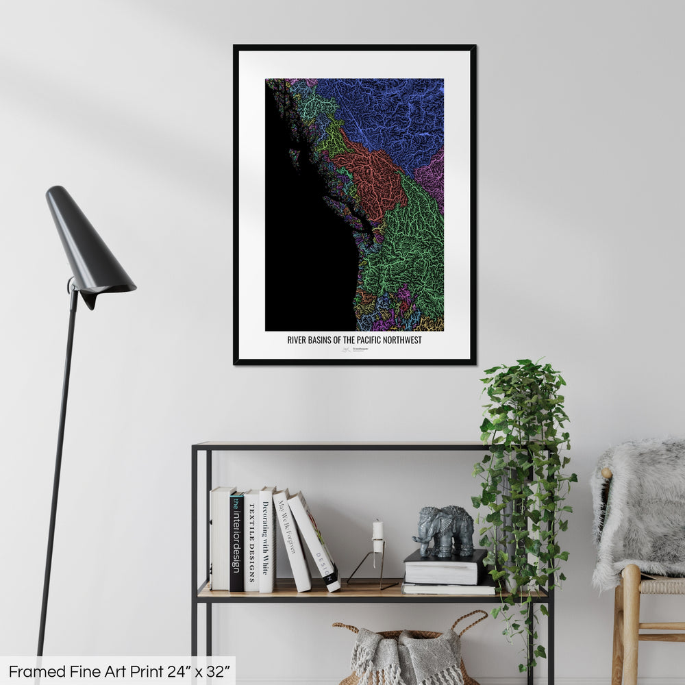 A colourful Grasshopper Geography map on a white wall, home decor in a living room. The map is in a black frame with a thick white mat. The title of the map is printed on the mat. The map hangs above a black shelf with plants and books on. A black lamp stands in the corner.