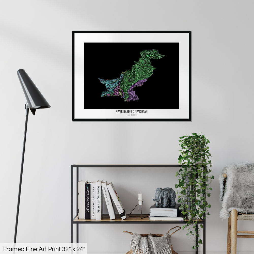 A colourful Grasshopper Geography map on a white wall, home decor in a living room. The map is in a black frame with a thick white mat. The title of the map is printed on the mat. The map hangs above a black shelf with plants and books on. A black lamp stands in the corner.