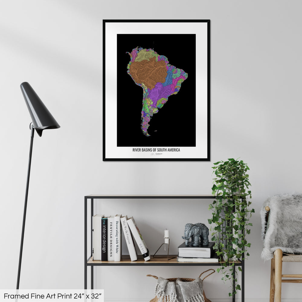 A colourful Grasshopper Geography map on a white wall, home decor in a living room. The map is in a black frame with a thick white mat. The title of the map is printed on the mat. The map hangs above a black shelf with plants and books on. A black lamp stands in the corner.