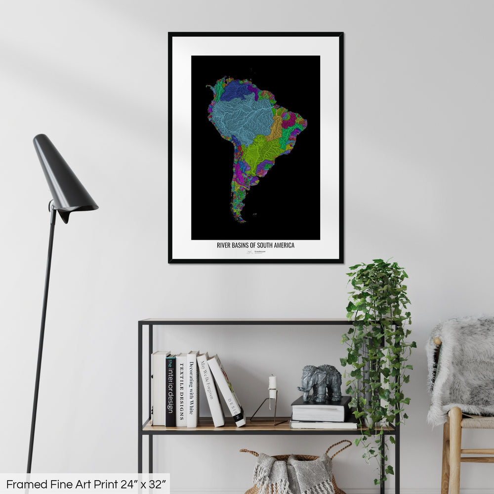 A colourful Grasshopper Geography map on a white wall, home decor in a living room. The map is in a black frame with a thick white mat. The title of the map is printed on the mat. The map hangs above a black shelf with plants and books on. A black lamp stands in the corner.