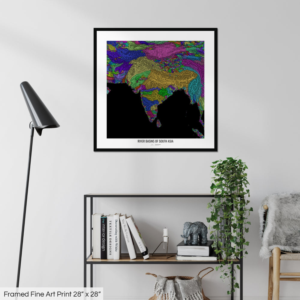 A colourful Grasshopper Geography map on a white wall, home decor in a living room. The map is in a black frame with a thick white mat. The title of the map is printed on the mat. The map hangs above a black shelf with plants and books on. A black lamp stands in the corner.