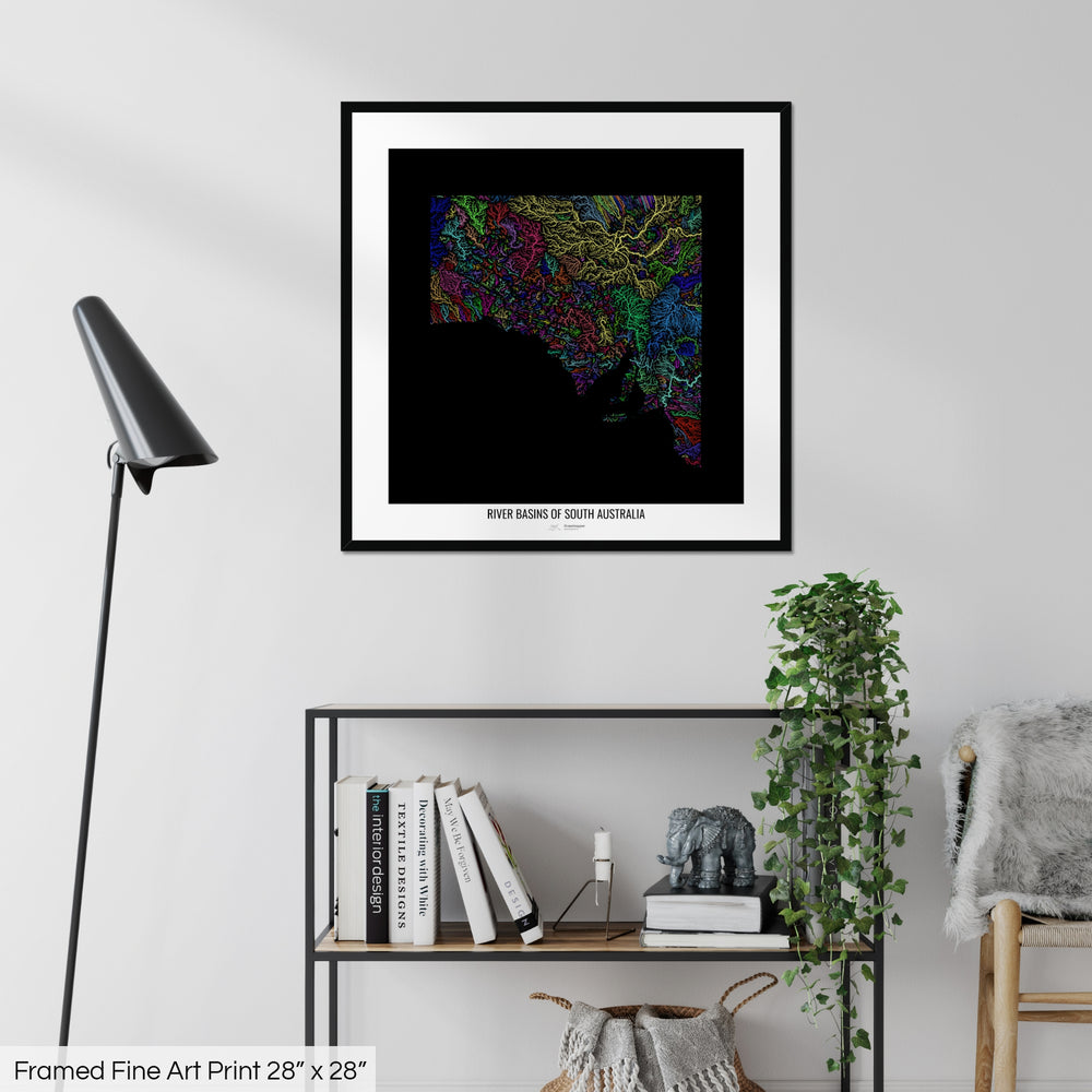 A colourful Grasshopper Geography map on a white wall, home decor in a living room. The map is in a black frame with a thick white mat. The title of the map is printed on the mat. The map hangs above a black shelf with plants and books on. A black lamp stands in the corner.