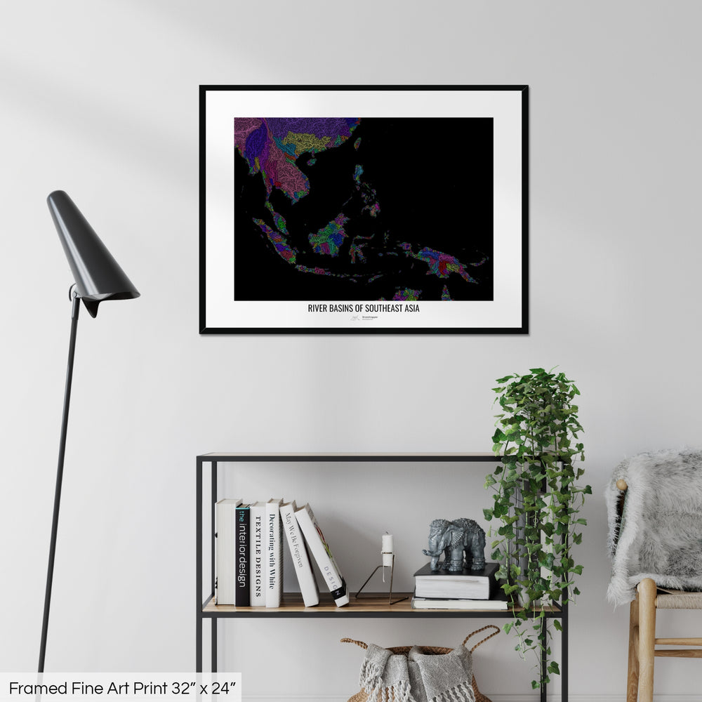 A colourful Grasshopper Geography map on a white wall, home decor in a living room. The map is in a black frame with a thick white mat. The title of the map is printed on the mat. The map hangs above a black shelf with plants and books on. A black lamp stands in the corner.