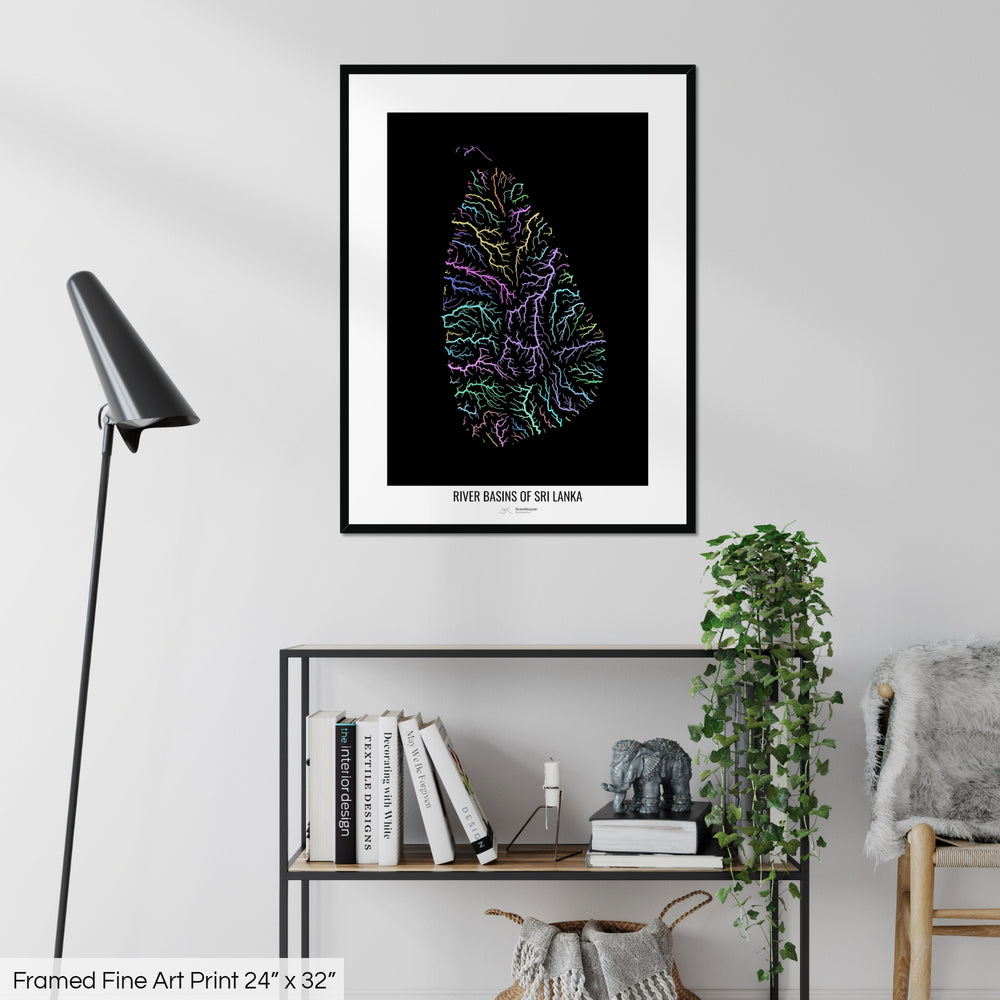 A colourful Grasshopper Geography map on a white wall, home decor in a living room. The map is in a black frame with a thick white mat. The title of the map is printed on the mat. The map hangs above a black shelf with plants and books on. A black lamp stands in the corner.