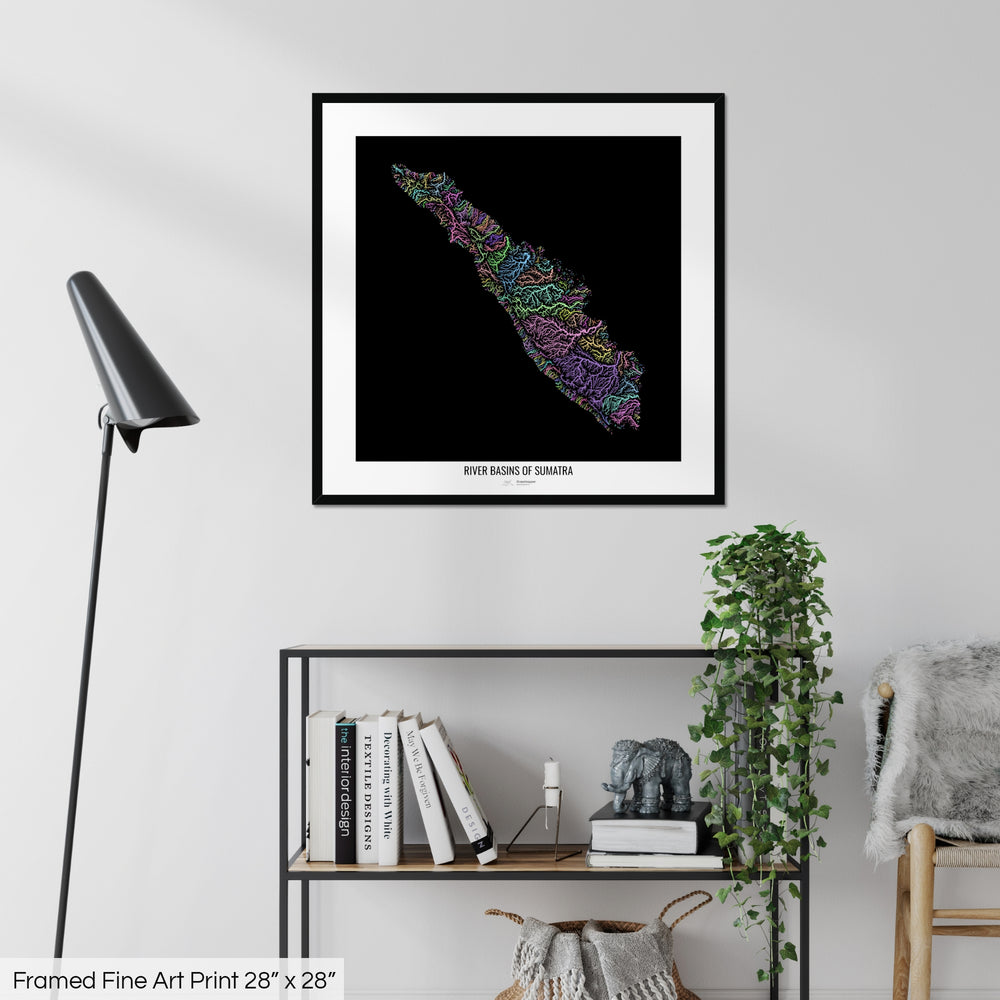 A colourful Grasshopper Geography map on a white wall, home decor in a living room. The map is in a black frame with a thick white mat. The title of the map is printed on the mat. The map hangs above a black shelf with plants and books on. A black lamp stands in the corner.
