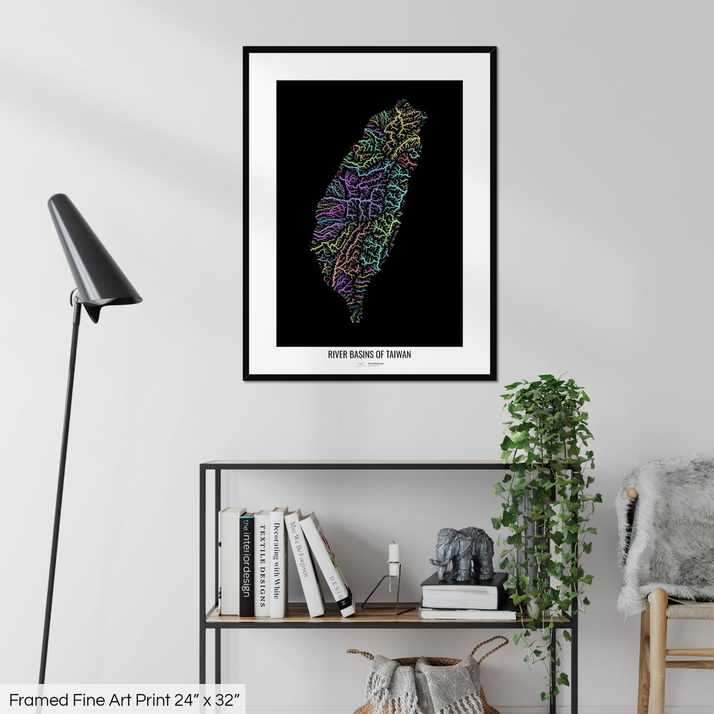 A colourful Grasshopper Geography map on a white wall, home decor in a living room. The map is in a black frame with a thick white mat. The title of the map is printed on the mat. The map hangs above a black shelf with plants and books on. A black lamp stands in the corner.