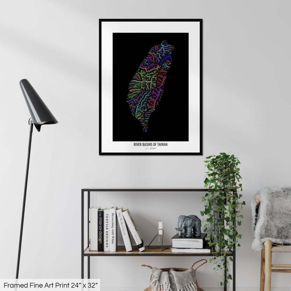 A colourful Grasshopper Geography map on a white wall, home decor in a living room. The map is in a black frame with a thick white mat. The title of the map is printed on the mat. The map hangs above a black shelf with plants and books on. A black lamp stands in the corner.