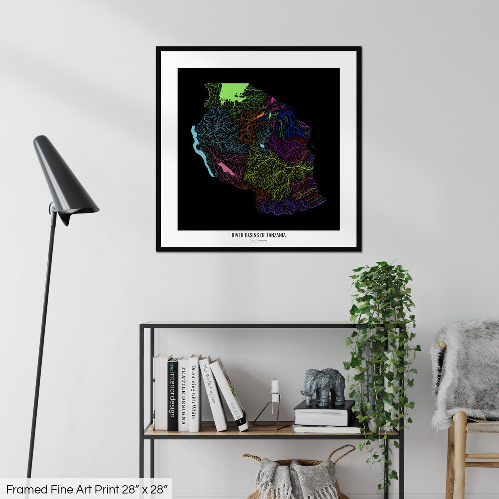 A colourful Grasshopper Geography map on a white wall, home decor in a living room. The map is in a black frame with a thick white mat. The title of the map is printed on the mat. The map hangs above a black shelf with plants and books on. A black lamp stands in the corner.