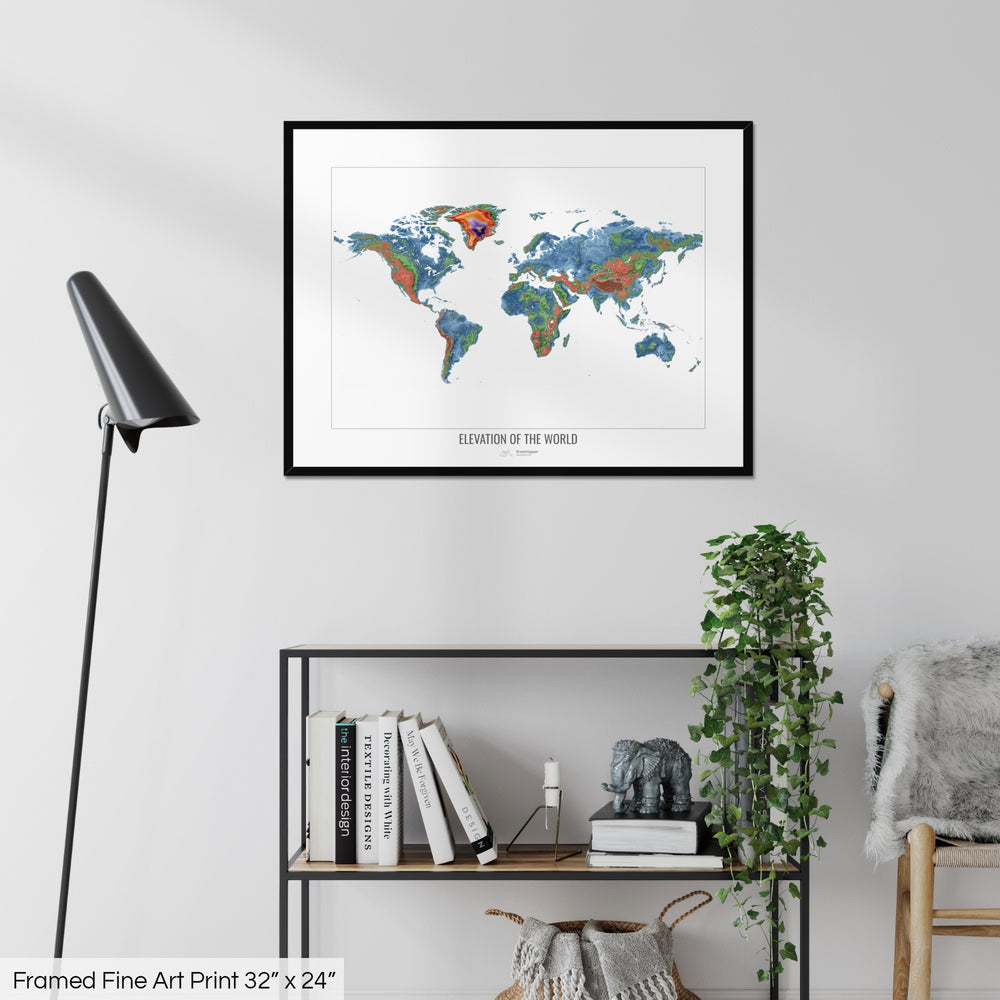 A colourful Grasshopper Geography map on a white wall, home decor in a living room. The map is in a black frame with a thick white mat. The title of the map is printed on the mat. The map hangs above a black shelf with plants and books on. A black lamp stands in the corner.
