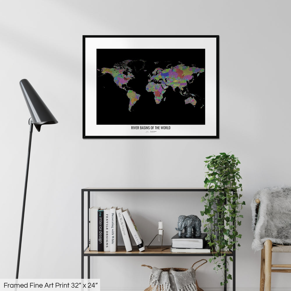 A colourful Grasshopper Geography map on a white wall, home decor in a living room. The map is in a black frame with a thick white mat. The title of the map is printed on the mat. The map hangs above a black shelf with plants and books on. A black lamp stands in the corner.