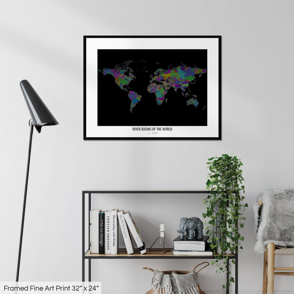 A colourful Grasshopper Geography map on a white wall, home decor in a living room. The map is in a black frame with a thick white mat. The title of the map is printed on the mat. The map hangs above a black shelf with plants and books on. A black lamp stands in the corner.