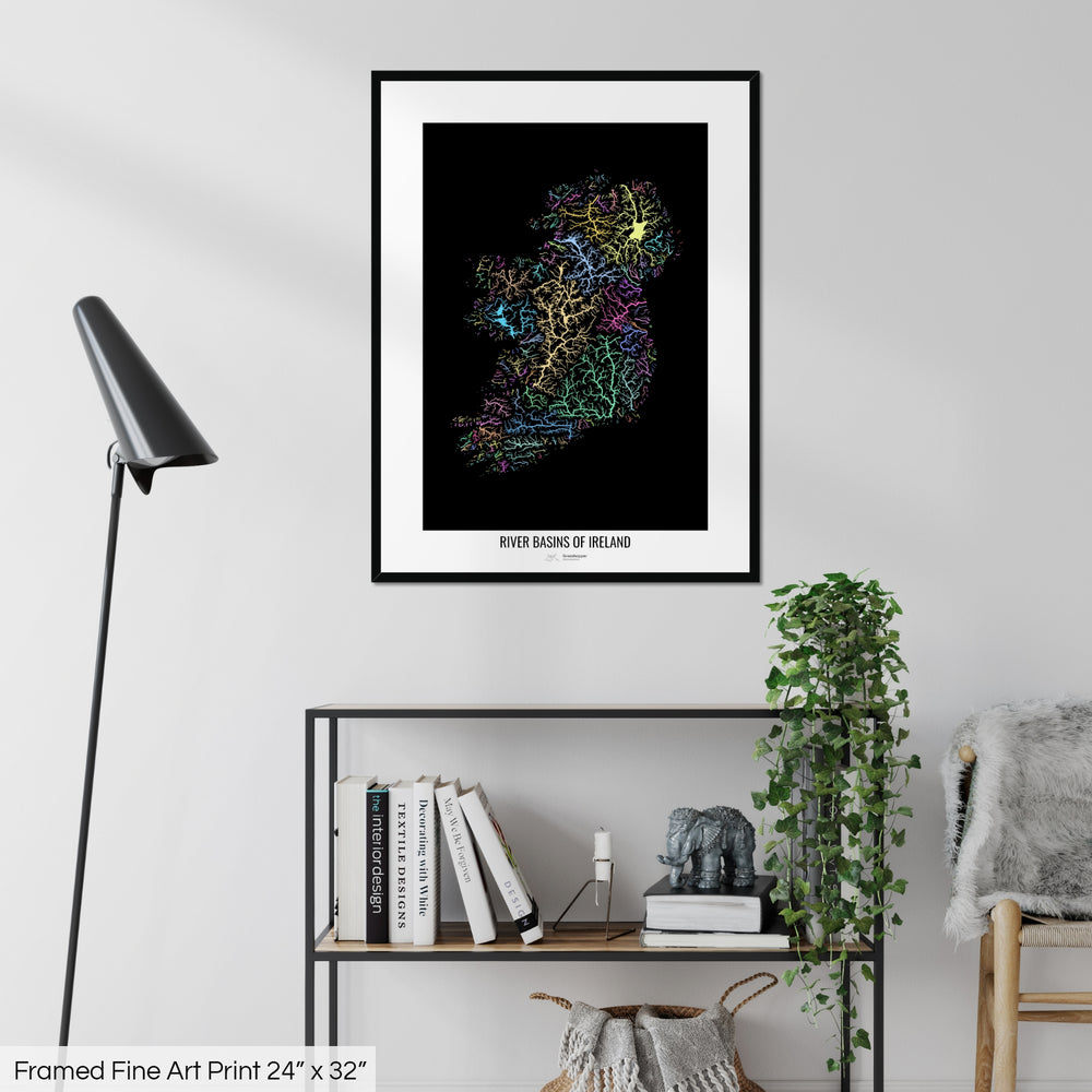 A colourful Grasshopper Geography map on a white wall, home decor in a living room. The map is in a black frame with a thick white mat. The title of the map is printed on the mat. The map hangs above a black shelf with plants and books on. A black lamp stands in the corner.
