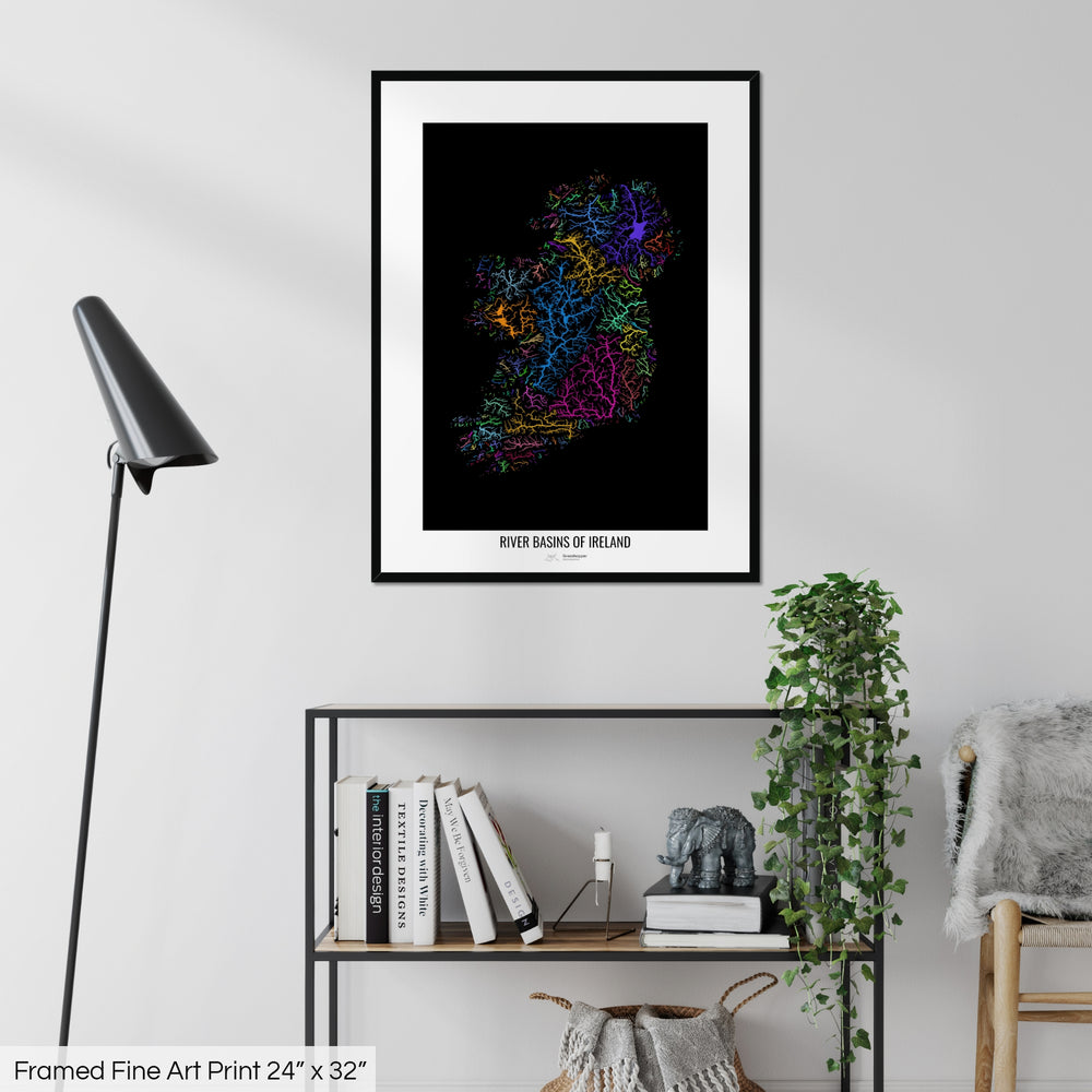 A colourful Grasshopper Geography map on a white wall, home decor in a living room. The map is in a black frame with a thick white mat. The title of the map is printed on the mat. The map hangs above a black shelf with plants and books on. A black lamp stands in the corner.