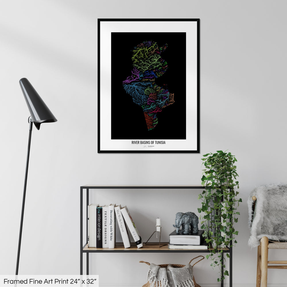 A colourful Grasshopper Geography map on a white wall, home decor in a living room. The map is in a black frame with a thick white mat. The title of the map is printed on the mat. The map hangs above a black shelf with plants and books on. A black lamp stands in the corner.