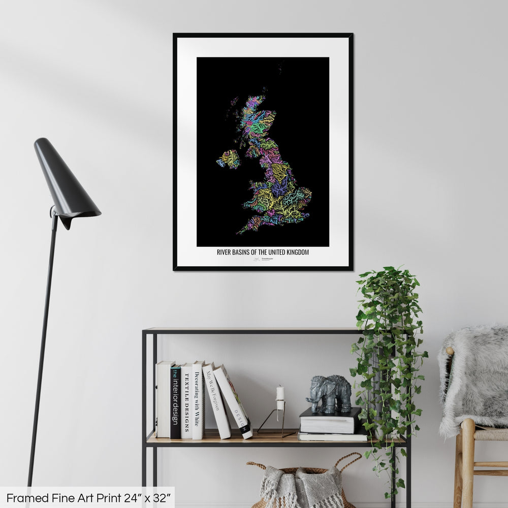 A colourful Grasshopper Geography map on a white wall, home decor in a living room. The map is in a black frame with a thick white mat. The title of the map is printed on the mat. The map hangs above a black shelf with plants and books on. A black lamp stands in the corner.