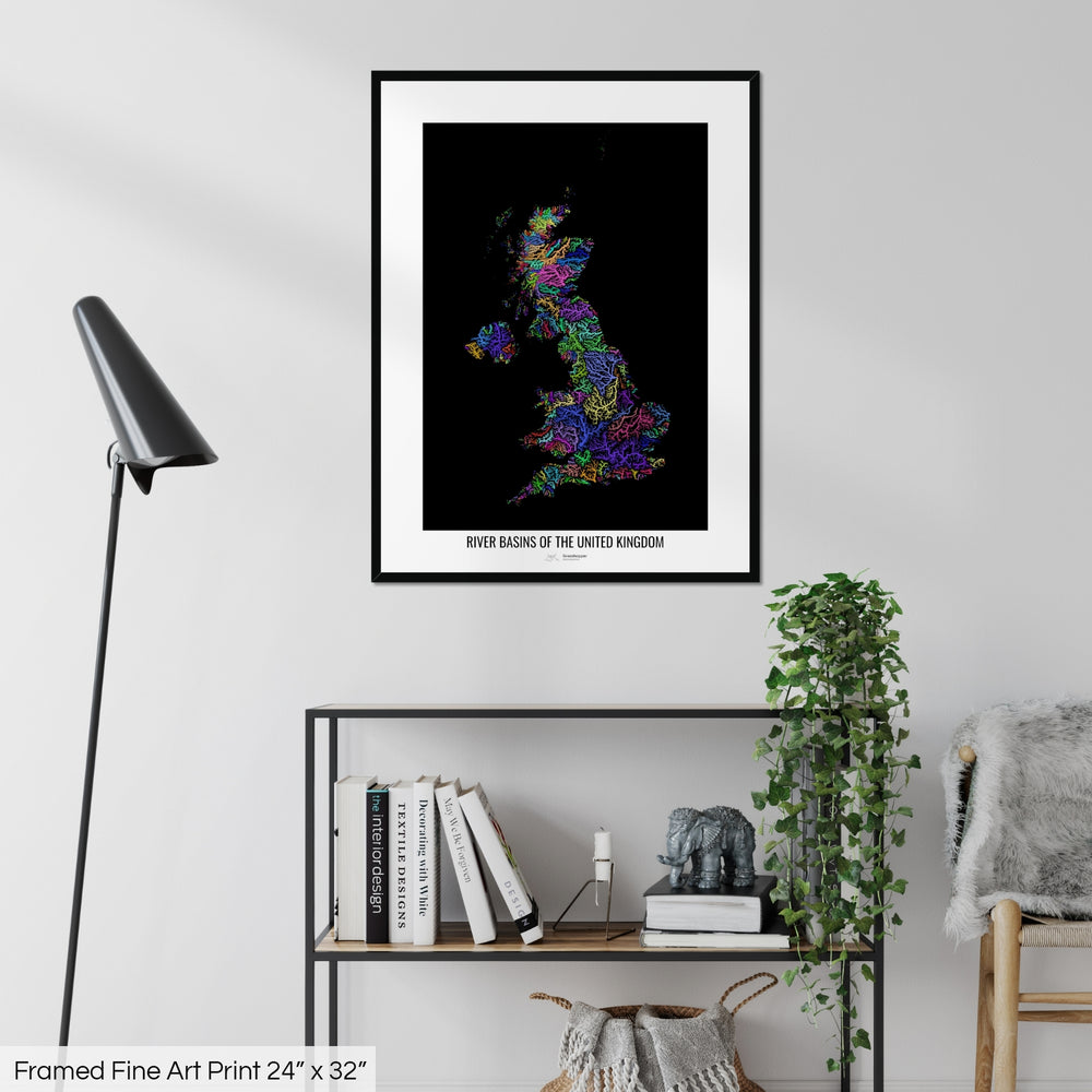 A colourful Grasshopper Geography map on a white wall, home decor in a living room. The map is in a black frame with a thick white mat. The title of the map is printed on the mat. The map hangs above a black shelf with plants and books on. A black lamp stands in the corner.