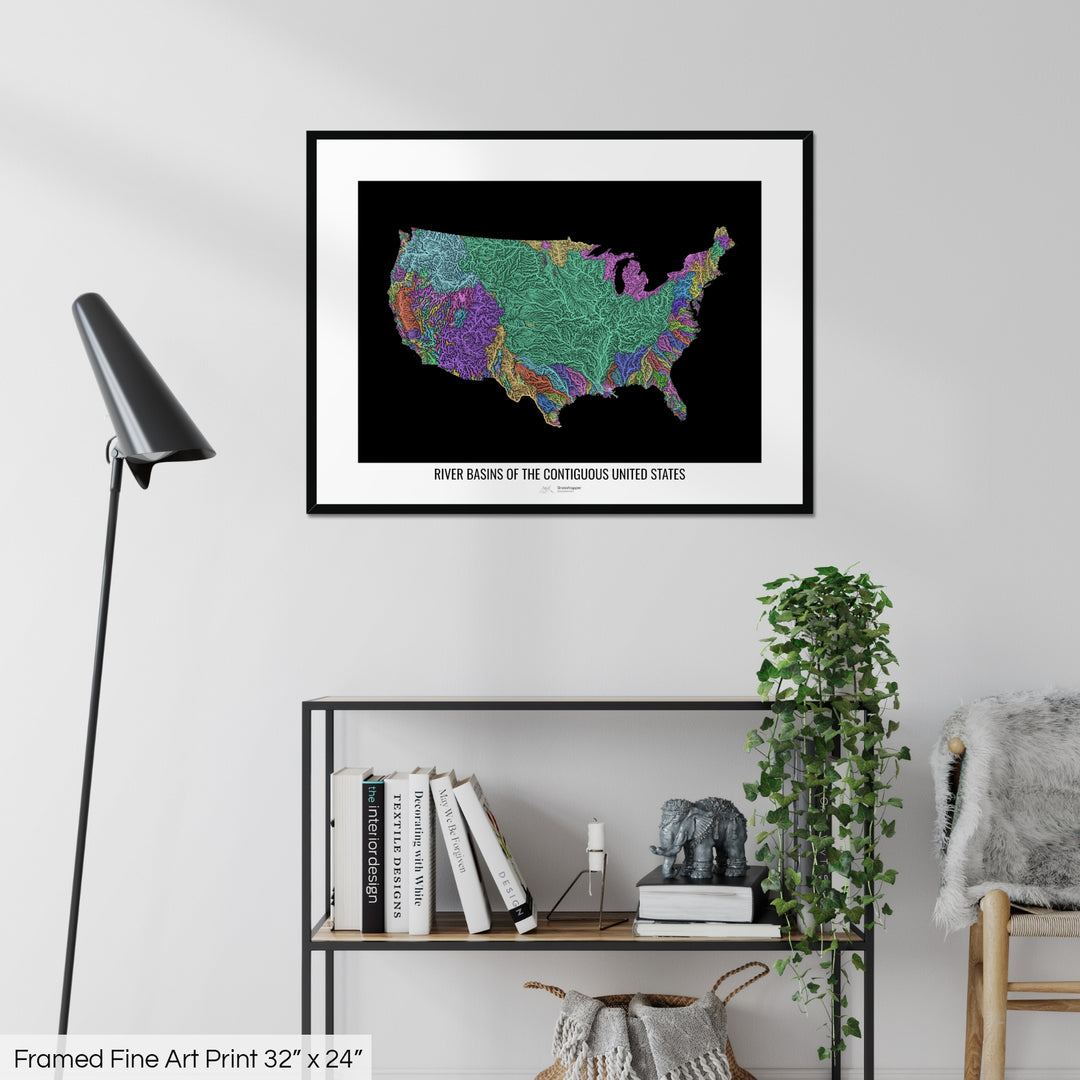 A colourful Grasshopper Geography map on a white wall, home decor in a living room. The map is in a black frame with a thick white mat. The title of the map is printed on the mat. The map hangs above a black shelf with plants and books on. A black lamp stands in the corner.