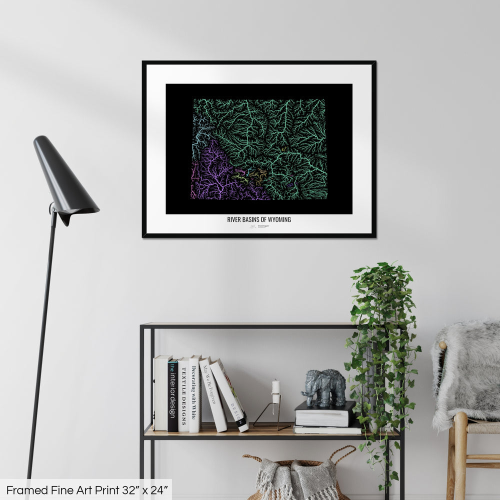 A colourful Grasshopper Geography map on a white wall, home decor in a living room. The map is in a black frame with a thick white mat. The title of the map is printed on the mat. The map hangs above a black shelf with plants and books on. A black lamp stands in the corner.
